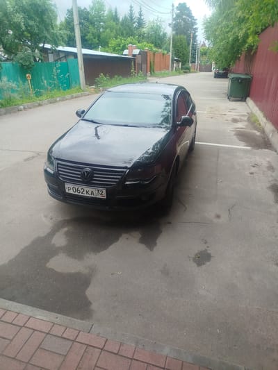 A black Volkswagen sedan is parked on an asphalt surface. The car is facing slightly to the left and its license plate is visible, reading "P062KA32". The car appears wet, suggesting recent rain. The environment is an outdoor urban setting, with a red corrugated metal fence on the right side of the frame and a green fence on the left. A large green dumpster is situated near the red fence. Trees and vegetation are visible in the background, indicating a suburban or semi-rural area. The ground also shows patches of wetness. The scene is consistent with a street or parking area in Nemchinovka, Russia.