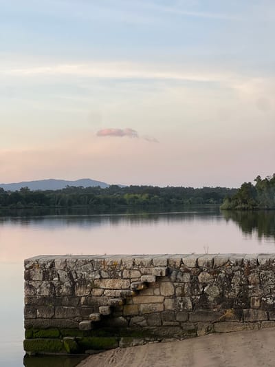 The image shows a landscape in Gondarém, Portugal. A body of water, likely a lake, reflects the sky and a densely forested shoreline. A stone structure, possibly a wall or bridge abutment, occupies the right side of the frame, exhibiting signs of age and weathering, including lichen growth. The sky is a gradient of pale blues and pinks, suggesting dawn or dusk.