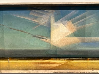 The image displays a framed painting titled *Sailboat on the Sea (No. 2)* by Fritz Glarner, dated 1923, exhibited at the Harvard Art Museums in Cambridge, Massachusetts, United States. The artwork, an oil painting, depicts a stylized marine landscape through geometric abstraction. The composition is horizontally divided into three primary sections: a sky, a horizon/sea, and a foreground. The upper section, representing the sky, consists of layered geometric planes in shades of light blue, teal, and beige, with an emphasis on diagonal and angular forms suggesting light refraction or clouds. A narrow, dark blue-grey horizontal band forms the horizon line. On the far left of this band, a small, upright white shape represents a sailboat. The bottom section, comprising approximately one-third of the canvas, depicts the foreground in varied golden and ochre tones, also rendered with geometric segments, suggesting a beach or sand dunes. The painting is enclosed within a light-colored, possibly wooden, frame, which is partially visible at the top and bottom edges of the photograph. The scene contains no human figures.