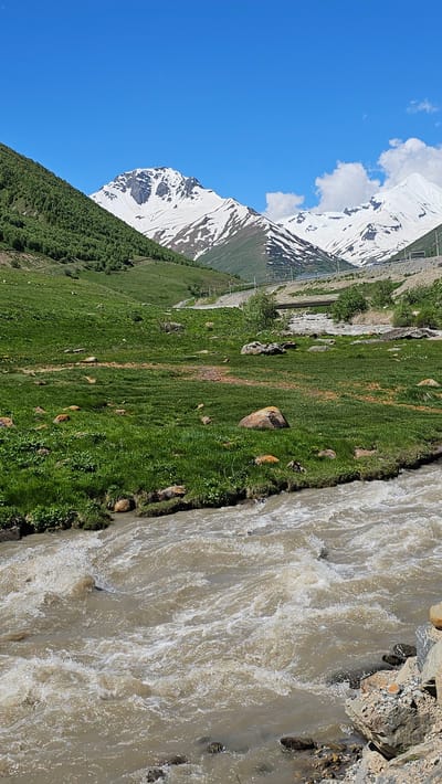 A vertical, eye-level shot captures a rushing, muddy river in the foreground, its turbulent waters filled with white foam and rapids. The river flows from the bottom right toward the bottom left of the frame. Scattered rocks line the immediate banks of the river, some submerged in the water.
Beyond the river, a lush green grassy meadow stretches uphill, dotted with various sized rocks. The meadow is bordered by a steep, tree-covered slope on the left, leading up to snow-capped mountains in the background. The peaks of these mountains are jagged and rugged, with patches of snow clinging to their upper reaches. A clear, bright blue sky with a few scattered white clouds dominates the upper portion of the image.
In the middle ground, a road curves through the landscape, with power lines and poles visible alongside it. A small concrete bridge spans a section of the landscape behind the road. The scene depicts a natural mountain landscape, likely in a rural or remote area, given the lack of prominent man-made structures beyond the road and bridge. The location is identified as Kamskho, Russia.