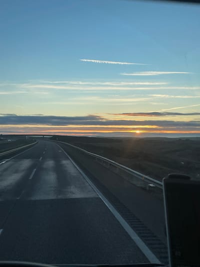 A road stretches into the distance, its asphalt surface reflecting light, flanked by barriers. The sky above is a canvas of blues and oranges, suggesting either a sunrise or sunset. Contrails streak across the blue expanse, while clouds in the distance obscure the horizon. The view is likely taken from within a vehicle, adding a sense of movement.