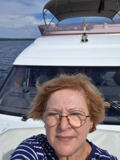 A woman with reddish-brown, wind-blown hair and round, gold-rimmed glasses is taking a selfie on a boat. She is wearing a blue and white striped shirt and a pearl earring. The boat is white and appears to be a motor yacht, with a canopy overhead. In the background, there is a calm body of water, a treeline on the horizon, and a cloudy sky. The location is the city of Shiryaevo, Russia.