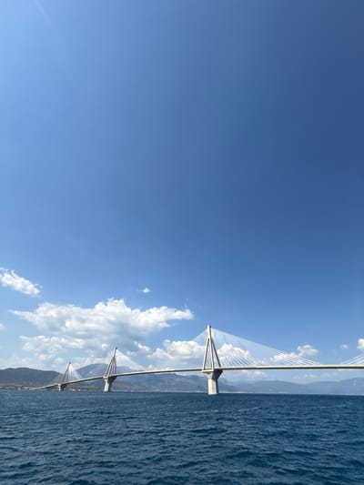 The image depicts a cable-stayed bridge spanning a body of water. The bridge is the primary object, with its tall pylons and numerous cables stretching across the frame. The water is a deep blue, with a textured surface indicative of waves or ripples. In the background, mountains rise up, some covered in greenery and others appearing rocky. The sky is a clear, bright blue with a few scattered white clouds, suggesting fair weather. The sun appears to be high in the sky, casting light and creating some highlights on the water and the bridge. The perspective is from the water level, looking towards the bridge and the distant landscape. No people are visible in the image. The scene conveys a sense of scale and engineering, highlighting the grandeur of the bridge and its natural surroundings. The Municipal Unit of Rio in Greece is known for the Rio-Antirrio Bridge, which fits this description.