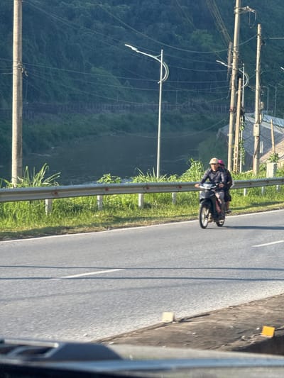 The image shows a road in Lao Cai Ward, Vietnam. The road is paved and has a white line marking the center. A person on a motorcycle is driving on the road. The road is bordered by a metal guardrail and green grass. Beyond the guardrail is a drop off with a forested hill. Power lines are visible extending from the left of the frame. A building roof can be seen in the upper left corner of the frame.
