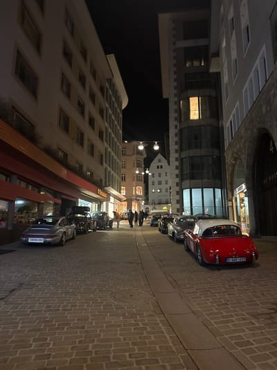 The image displays a nighttime street scene in Sankt Moritz, Switzerland, specifically a cobblestone street near the Rathaus (City Hall), which is visible in the background. Multi-story buildings line both sides of the street, illuminated by various artificial light sources.

On the left, a light-colored, multi-story building features numerous windows and commercial storefronts at ground level; one sign clearly reads "SPIRKENBERG." On the right, the architecture varies, including a grey-facade building, a modern glass-fronted structure with a brightly lit interior revealing a chandelier, and an older building distinguished by a large stone archway.

A collection of classic automobiles is parked along both sides of the street. On the left, from the foreground, a silver Porsche 911 (Swiss license plate "ZH 901 109") is parked, followed by a dark vintage convertible with luggage secured on its boot, and other classic vehicles extending down the street. On the right, a red vintage convertible sports car with a white hardtop, marked with the number "21" and bearing a British license plate "O AGF 037," is parked. Behind it, a grey vintage convertible is visible, succeeded by additional classic cars.

Approximately 6-8 pedestrians are observable walking along the street in the middle distance. One individual, wearing an orange jacket, stands near a dark vintage car parked on the left side of the street. The street surface is composed of rectangular cobblestones, with a distinct narrow drainage channel running along the center-right.