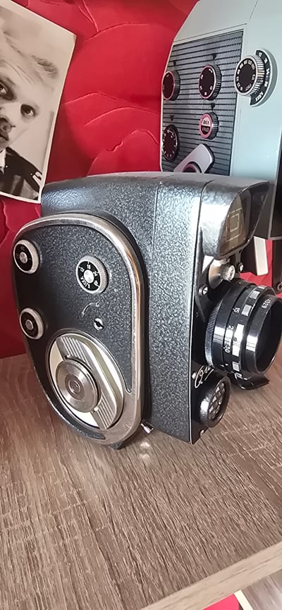This image shows a vintage camera, possibly a Super 8 film camera, placed on a wooden surface. The camera is gray and has a textured finish. It features several dials and knobs, some with visible inscriptions. A prominent lens is attached to the front. Next to the camera, a black and white photograph of a person, potentially an actor or celebrity, is partially visible. The background includes a red, textured surface, possibly upholstery, suggesting the camera is on furniture. The setting appears to be indoors, with artificial lighting. The camera has various settings labeled, including numbers and Cyrillic text which appears to say "Е.А." and "ГОСТ". Another dial has "15", "2", "30" and "4329" written on it, as well as "V.I.R". The overall mood is one of nostalgia and appreciation for vintage technology. The camera itself is the primary subject, with the photograph adding a layer of historical context. The image is taken from a high angle, looking down at the camera.