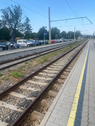 The image depicts a railway station platform in Eisenstadt, Austria. The scene is dominated by train tracks running parallel to the platform, which is paved with gray interlocking concrete blocks and marked with yellow and dark blue safety lines. On the left side of the tracks, a concrete retaining wall separates the tracks from a parking area where several cars and a white van are parked. Behind the parking lot, there are trees and utility poles with overhead wires for the electric trains. The sky is clear and blue. There are no people visible in the image. The overall impression is one of a functional, operational railway station in a suburban or semi-urban setting.