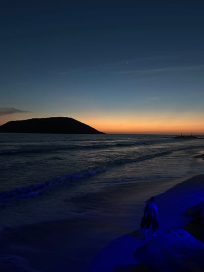 The image captures a beach scene in Mazatlán, Mexico at sunset. The sky transitions from dark blue to orange. A dark mountain or hill is silhouetted against the lighter part of the sky. The ocean's waves reach the shore, with bluish hues reflecting the fading light. A person stands near some dark rocks, likely along the shoreline. The overall scene is dimly lit, creating a somber and tranquil atmosphere.