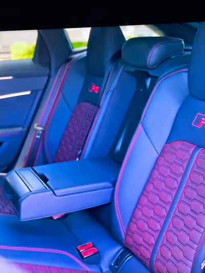 The image is a close-up shot of the interior of a car, specifically focusing on the rear seats. The seats are upholstered in dark blue leather with prominent red stitching outlining the contours and detailing. The seat inserts feature a textured hexagonal pattern in a deep purple or magenta color, also accented by red stitching. A central armrest, made of the same dark blue material, is folded down between the seats. A red seatbelt buckle is visible on the lower right portion of the image. Red "RS" logos are subtly embossed on the seatbacks. The background is blurred, showing hints of green foliage and a light-colored building, suggesting the car is parked outdoors. There are no people visible in the image. The scene is likely a professional photograph intended to showcase the car's interior design and features. The image does not provide any information about the city of Bezden, Bulgaria.