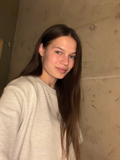 A young woman with long, dark brown hair is taking a selfie in a dimly lit room. She is wearing a light-colored crewneck sweatshirt. The background appears to be a concrete wall with some discoloration and markings. There are some small, possibly decorative, objects hanging on a wire against the wall. The woman is looking directly at the camera and smiling slightly. The lighting suggests an indoor environment, possibly a home or a less polished commercial space. The city of Volochysk, Ukraine is not depicted in the image, nor is any specific urban environment suggested by the background.