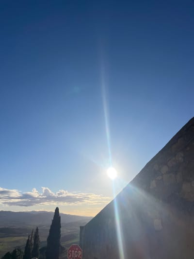 This image captures a vibrant, sun-drenched landscape, likely from an elevated perspective overlooking the rolling hills of Pienza, Italy. The dominant feature is the expansive, clear blue sky, filling the upper portion of the frame. A brilliant sun, positioned high in the sky, emits a strong lens flare that streaks across the upper middle, indicating a bright midday or early afternoon.

Near the horizon on the right side, soft white clouds drift above a vast landscape of undulating green fields and distant hills, characteristic of the Tuscan countryside. Several dark green, conical trees, resembling cypress trees, are visible on the far right, partially silhouetted against the bright backdrop. Along the top right edge, a sturdy stone or concrete wall is visible, suggesting a viewpoint or structure.

In the bottom right corner, a red octagonal stop sign with the word "STOP" clearly visible in white letters is partially obscured by foliage or the frame's edge. No specific activity or people are present. The overall impression is one of serene natural beauty under a radiant, clear sky.