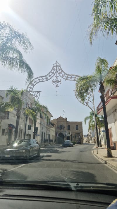 The image shows a street in Maruggio, Italy, with buildings on either side and palm trees. There is a decorative archway spanning the street, adorned with lights and intricate patterns. A dark-colored Audi A6 Avant is parked on the left side of the road, facing forward. In the distance, a light blue car is visible. The sky is clear and bright, with the sun casting a glare on the scene. The architecture suggests a Mediterranean setting, and the overall atmosphere is that of a sunny day in a town square or a main thoroughfare.