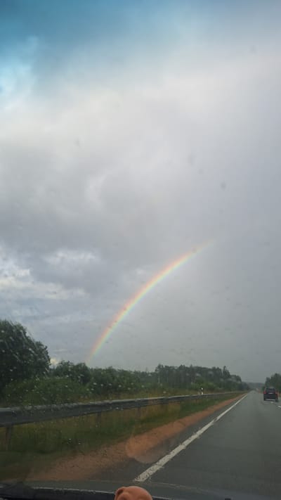 The image depicts a scene from a car driving on a highway, likely in or near Ogresgals, Latvia. A rainbow arches across a cloudy sky, partially obscured by rain droplets on the windshield. The highway stretches into the distance, with a dark-colored SUV visible on the right lane. A metal guardrail separates the highway from a grassy median and a line of trees and foliage on the left side. The overall atmosphere suggests a rainy or post-rain day. The bottom of the frame shows the dashboard and a hint of a person's hand, indicating the perspective of the driver or passenger.