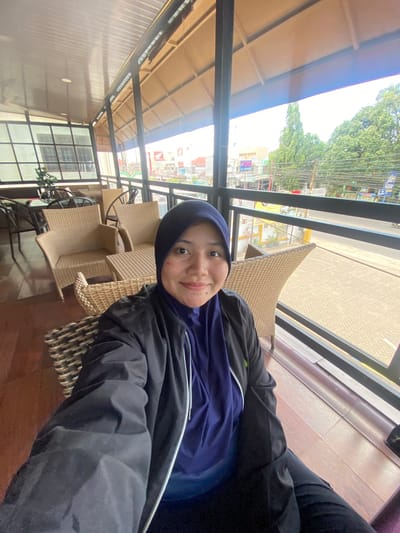 A young woman wearing a blue hijab and a black jacket is sitting on a woven chair in an outdoor cafe in Keraton, Indonesia. The cafe has a covered patio with a corrugated metal roof and large windows that offer a view of a street lined with trees and buildings. The woman is smiling at the camera and holding a phone, suggesting she is taking a selfie. The cafe furniture is also made of woven material, including chairs and tables. In the background, there are more tables and chairs, as well as some greenery. The overall atmosphere appears relaxed and casual.