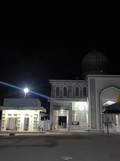 At night, a brightly lit mosque with a patterned dome is visible in Keudealue Ie Puteh, Indonesia. An adjacent smaller building with open stalls and a solitary chair is also illuminated. A sign is present near the mosque's entrance. The ground is dark and appears to be a paved area. The overall environment is dark.