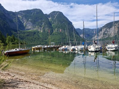 This image depicts a tranquil scene at the harbor in Obertraun, Austria. Several sailboats are docked along a wooden pier. The water is clear, with the sandy bottom visible in the foreground. Lush green mountains form a majestic backdrop to the scene, with scattered clouds in the bright blue sky. The sun is shining, casting reflections of the boats and the mountains onto the water. The environment is natural and serene, with no people visible in the frame.
