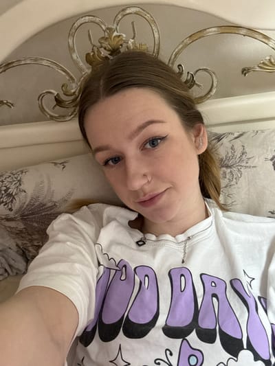 A single young adult female is depicted in a close-up, selfie-style image. She possesses light skin, light brown hair, and blue eyes, accented with winged black eyeliner. A small silver hoop nose ring is visible on her left nostril, and multiple small, clear-stoned earrings are present in her right earlobe, while a single stud is in her left. She wears a white t-shirt with a purple and black graphic design that includes the phrase "GOOD DAY" and small stars. A thin silver-colored necklace with a dark rectangular charm and a small teardrop-shaped pendant is around her neck. She is positioned against a light-colored pillow with a dark botanical pattern. Behind her head, an ornate light-colored headboard with gold-painted scrollwork is visible. The scene is indoors, consistent with a bedroom, located in the city of Tlustenkhabl, Russia.