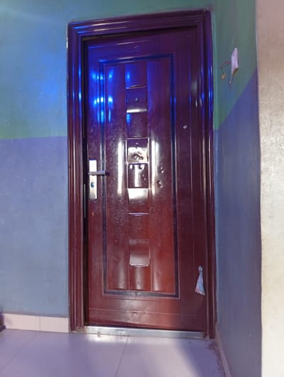 A maroon metal door with a silver handle is set in a doorway. The door is paneled with rectangular indentations. A small piece of patterned fabric is tied to the doorframe. The wall to the left of the door is painted in two horizontal sections: a lighter blue at the bottom and a darker green at the top. The wall to the right of the door is also painted in two horizontal sections: a darker blue at the bottom and a lighter beige at the top. A white electrical outlet with exposed red and green wires is visible on the green section of the wall to the right of the door. The floor in front of the door is tiled with white squares. The scene is illuminated with cool blue light, possibly from an external source. This image was taken in Olambe, Nigeria.