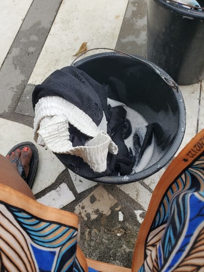 The scene, captured in Isunba, Nigeria, presents a high-angle view of a person's lower body and laundry implements on a tiled outdoor surface. The individual wears black flip-flops, displaying red nail polish on their toenails, and patterned trousers featuring blue, brown, and tan motifs. A black plastic bucket, partially filled with soapy water and containing black and light-colored pleated clothing, occupies the central foreground. A small, light-brown bird rests on the rim of this bucket. A second, similar black bucket is partially visible behind and to the right of the first. The ground consists of light-toned tiles with darker grout lines and wet, worn areas.