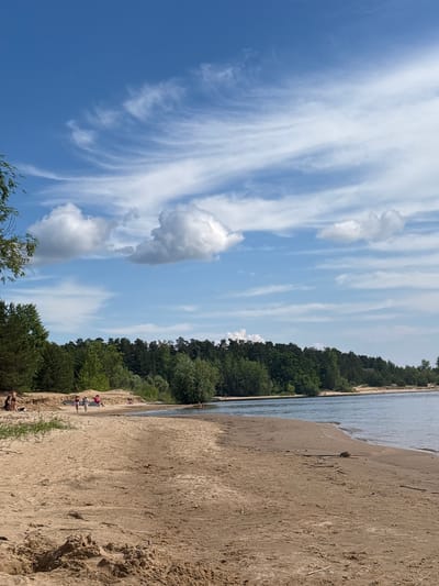 A wide shot of a sandy beach in Kzyl-Bairak, Russia, reveals a calm body of water on the right side and a dense forest in the background. Several people are scattered across the beach, some walking, some sitting, and others playing near the water's edge. The sky is a vibrant blue with streaks of white clouds. The sand is light brown and appears slightly damp in some areas closer to the water. Trees, predominantly coniferous, form a dark green line along the horizon, with some deciduous trees interspersed. The overall scene depicts a leisurely day at a natural outdoor setting.