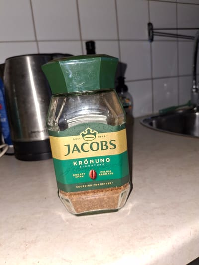 A jar of Jacobs Krönung instant coffee sits on a speckled countertop in a kitchen. The jar is mostly full of coffee granules. The label on the jar is gold and green with the Jacobs logo and "Krönung Signature" printed on it. To the left of the jar is a stainless steel kettle. In the background, there are white tiled walls and a faucet. The scene is illuminated by overhead lighting. The location of Åsgårdstrand, Norway is not discernible from the image.