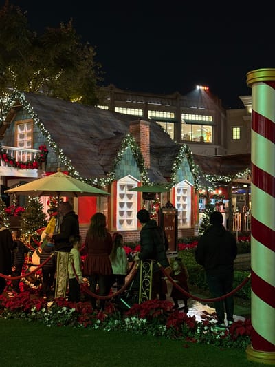 A group of people are lined up behind a red velvet rope near a Christmas-decorated building. The building is covered in lights and garlands, and has a red and white striped pole next to it. There is a light in the sky behind the building. The scene is festive and looks like it is from a Christmas-themed event or attraction.