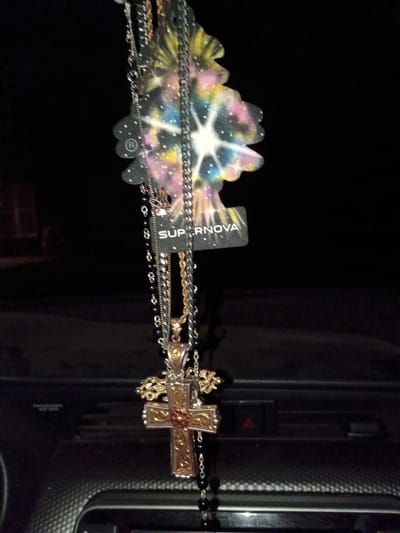 The image shows a cross necklace and other necklaces dangling from a Supernova brand tag. The background appears to be the interior of a car, specifically the dashboard area. The location is potentially Clarksville, United States of America.