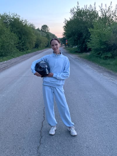 A person stands on a paved road in Doma 45 Kilometr, Russia. The person wears a light blue tracksuit and white sneakers, holding a black motorcycle helmet. Trees line the left side of the road, and the sky is visible on the horizon. A speed limit sign and a stop sign are visible in the background to the left. The road appears to be asphalt.