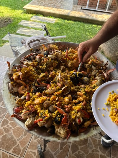 A large paella, filled with seafood and rice, is being served. The paella includes crabs, mussels, clams, and shrimp. A hand is using a spoon to serve a portion of the paella onto a white plate. The paella is in a large metal pan supported by a stand. The scene appears to be outdoors, with grass and tiled patio flooring visible. The location could be San Francisco Lachigoló, Mexico. A building is visible in the background.