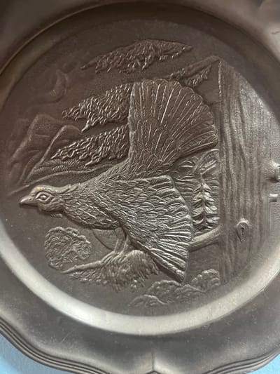 This is a detailed photograph of a circular relief plaque, likely made of pewter or a similar metal, featuring an embossed scene of a grouse in flight amidst a mountainous, forested landscape. The plaque is oval-shaped with a raised rim.
The central subject is a large grouse, depicted in mid-flight with its wings spread and fanned out, revealing intricate feather details. The bird's head is turned to the left, with its eye and beak clearly defined. It appears to be perched on a branch or a stylized tree.
The background depicts a textured mountainous terrain with stylized trees and foliage. To the right, a large tree trunk is prominently featured, with bark textures and a knot or branch visible. In the upper left corner, there's a suggestion of a crescent moon or a curved mountain ridge. The overall impression is one of a natural, wilderness setting.
The lighting emphasizes the raised details of the relief, creating shadows that add depth and dimension to the scene. The plaque exhibits some minor surface wear and subtle scratches consistent with age and handling.
This object is likely decorative and could be a souvenir, a commemorative item, or a piece of artistic craftsmanship. Without further context, its specific location in Schwäbisch Hall, Germany, cannot be determined from the image alone.