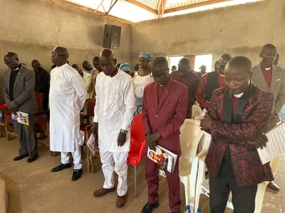 The image depicts a group of men, likely clergy, standing in a sparsely furnished room that appears to be a place of worship. The setting is rustic, with a corrugated metal roof supported by wooden beams and unfinished concrete walls. The floor is a simple concrete slab. Several men are dressed in clerical attire, including white cassocks and robes, while others wear suits and robes. They appear to be standing in prayer or during a service, with their heads bowed and eyes closed.

In the foreground, several men are prominently featured. One man on the left wears a gray suit and glasses. Next to him, a man in a pristine white traditional robe and trousers stands with his eyes closed. To his right, another man in a maroon suit with a dark patterned vest and tie is also in prayer. Further to the right, a man in a distinctive black and red patterned suit and black trousers is visible.

Some of the men are holding what appear to be programs or bulletins. One bulletin visible in the foreground has a picture of a man in clerical attire and text that includes "REV. BOLUS ALI". Another bulletin in the hands of the man in the gray suit displays a collage of images.

The lighting suggests it is daytime, possibly late morning or early afternoon, with natural light filtering in from unseen windows or openings, augmented by artificial lighting from what might be speakers mounted on the wall. The overall mood conveyed is one of solemnity and devotion. The context of Kufana, Nigeria, suggests this scene is likely a church service or religious gathering in that region.