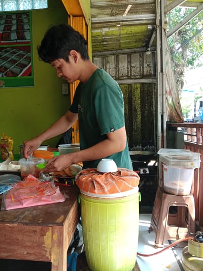 This image captures a man preparing food at a rustic street food stall in Sekopek Wetan, Indonesia, under bright daytime lighting. The scene suggests a bustling, local eatery.
The central subject is a man, likely in his 20s or 30s, seen in profile from the chest up, facing left. He has dark, medium-length hair and is wearing a dark green t-shirt with a small white "dlx" logo on the left sleeve. His hands are actively engaged in food preparation, placed over a clear plastic container holding a red or orange sauce, while he holds a small green dish.
The foreground features a worn wooden counter, upon which various items are arranged. These include several clear plastic containers with lids (some open), a yellow pouch labeled "MINYAK SUNNAH" (cooking oil), and multiple plastic bags, including a pink one with red text. Beside the counter, a light green plastic barrel, possibly a cooler or storage container, is draped with an orange-patterned cloth and topped with a small white bowl. To the right, a brown plastic stool holds another large clear plastic container. On the ground, a yellow gas cylinder with a red hose connects to a small gas stove, indicating active cooking.
The background shows the structure of the stall. The left wall is painted bright green and features two framed posters depicting various food items in square boxes. The upper sections of the stall and roof are constructed from corrugated metal, showing signs of rust and wear. An exposed light bulb hangs from the roof. Bright sunlight streams in from the right, illuminating parts of the scene and casting shadows. Beyond the stall's opening on the right, green foliage, trees, and a partial view of a vehicle are visible, confirming the outdoor setting. The overall impression is of a functional and well-used food vendor preparing meals.