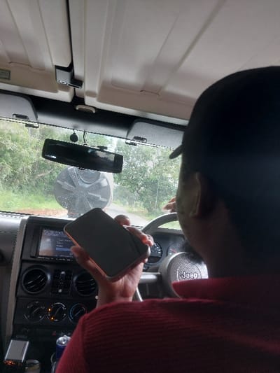 The image depicts the interior of a Jeep vehicle in Kisanga, Tanzania, viewed from a rear-passenger perspective. A person, identified as the driver, is observed from behind, wearing a dark cap and a red, textured collared shirt. The individual's right hand holds a dark-colored smartphone, equipped with a light-colored protective case, displaying a dark, unlit screen. The vehicle's dashboard, steering wheel bearing the "Jeep" logo, infotainment system, air vents, and climate controls are visible. Through the windshield, an outdoor scene unfolds, featuring a paved road curving to the left, bordered by green foliage and trees. Prominently positioned in the background, near the roadside, is a large, dark grey or black corrugated water storage tank.