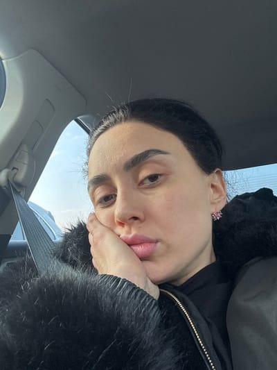 A selfie shows a person resting their face on their hand, wearing a black furry jacket and a pink earring. The person appears to be inside a car, with the car's ceiling and a seatbelt visible in the frame. Outside the car's window, a glimpse of the sky can be seen. The overall perspective is angled, looking upward towards the person.