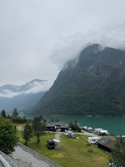 The image depicts a campsite located in Olden, Norway, with a dramatic mountain landscape and a vibrant turquoise lake as the backdrop. The campsite features several static caravans, tents, and recreational vehicles scattered across a grassy area. A dark-colored van is parked with an awning extended, and picnic tables are visible nearby. Small, dark-colored cabins are also present on the property. A road runs along the left side of the image, separated from the campsite by a metal barrier. The surrounding mountains are covered in dense green forest, with some peaks shrouded in clouds. The sky is overcast and grey, contributing to a serene and somewhat moody atmosphere. The water of the lake is a striking teal color. There are no visible people in the scene.