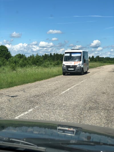 A white passenger van with the text "ФЕНИКС 240 240" on its side and front is driving on a paved road. The license plate on the front of the van reads "А831АР 67". The van has orange and white stripes on its front. The road is flanked by a grassy field on the left and a line of trees in the background. The sky is blue with scattered white clouds. The image appears to be taken from inside another vehicle, as the hood and windshield wipers are visible in the foreground. The location is Izdeshkovo, Russia.
