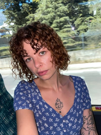 A woman with curly brown hair and green eyes is captured in a close-up selfie. She is wearing a blue dress with a white floral pattern and has tattoos on her chest and arm. The background appears to be a blurry landscape of trees and buildings, suggesting she might be traveling by vehicle. The lighting is bright, casting a slight sheen on her skin.