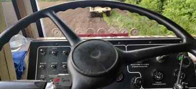 The image shows the interior of a heavy-duty vehicle, likely agricultural or construction equipment, from the driver's perspective. The steering wheel dominates the foreground. The dashboard features various gauges, switches, and indicators, including controls for lights, work lights, a beacon, a heater, a wiper, differential lock, and shutdown override. There's also a "SELECT PARK TO ENABLE STARTER OPERATION" notice, along with gauges for oil temperature, engine oil pressure, and a digital voltage display. A water bottle and a blue cloth are visible to the left.
Looking through the windshield, the view reveals a tilled field with rows of young plants. In the distance, another vehicle or piece of equipment is driving along the field. The surrounding environment includes vegetation on the right side of the view, suggesting a rural or agricultural setting.
Given the text "Grote" on a component and the fact that heavy machinery is in use, there's nothing that supports the claim that the location is in the city of Kovalivs'ke, Ukraine.