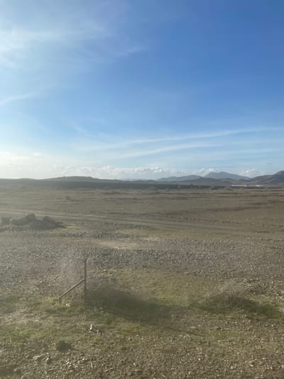 The image depicts a wide, open, and somewhat arid landscape under a clear blue sky, likely in Jbilate, Morocco. The scene is dominated by a vast expanse of ground in the foreground and mid-ground, transitioning to distant, hazy mountains or hills along the horizon.
The sky occupies the upper portion of the frame, showcasing a vibrant blue color with streaks of wispy white clouds, characteristic of high-altitude cirrus clouds. The light appears bright, suggesting either morning or late afternoon.
The ground comprises predominantly gravelly or rocky soil, interspersed with patches of sparse, dry green and brown vegetation. A faint, lighter-colored track or road is visible winding through the terrain, suggesting vehicular passage. In the immediate foreground, towards the right side of the image, a single, slender wooden or metal post is seen, leaning slightly. Its purpose is not explicitly clear, but it could be a marker or part of a simple, broken fence.
No people or significant activities are depicted, giving the scene a tranquil and uninhabited feel. The overall impression is one of natural ruggedness and vastness. There is no visible text in the image. The general atmosphere suggests dry, clear weather conditions.