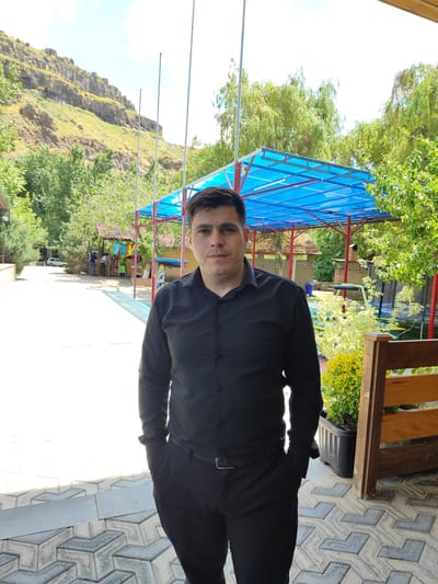 A man stands outdoors, facing the camera. He has dark hair and is wearing a black collared shirt and black pants. His left hand is in his pocket. The background features a rocky hillside, trees, and a structure with a blue roof. The ground is paved with interlocking tiles. The scene appears to be in a park or recreational area, potentially in the city of Karbi, Armenia, given the landscape features.