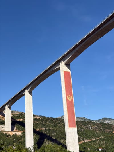 The image displays a large concrete viaduct, located in Gornje Mrke, Montenegro, viewed from a low-angle perspective looking upwards. The structure consists of multiple tall, slender concrete piers supporting a broad, flat bridge deck. One prominent pier, positioned towards the center-right of the frame, features a large, vertically oriented rectangular banner painted in red with a golden border. Centered on this red banner is the golden Coat of Arms of Montenegro, depicting a double-headed eagle clutching an orb and scepter, with a lion on its chest shield. The top edge of the bridge deck is lined with a series of white, spike-like elements. The viaduct spans across a valley characterized by steep, undulating hillsides densely covered in various green vegetation and trees. The background shows distant mountains under a clear, vibrant blue sky. Distinct shadows are cast by the bridge piers and the terrain, indicating bright sunlight. A small white building with a red roof is discernible at the base of a distant hill on the lower right side. There are no people, vehicles, or animals visible in the scene.