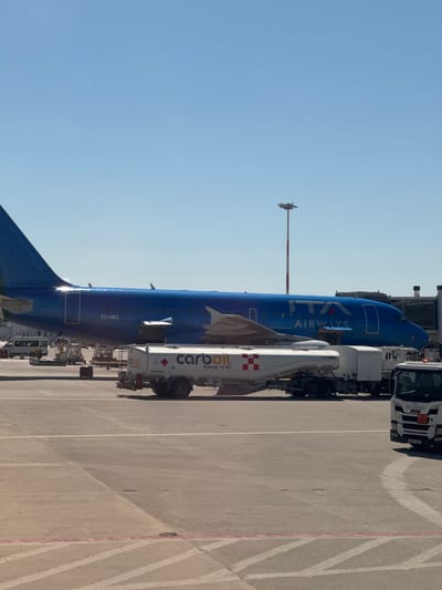 The image depicts an ITA Airways airplane being refueled on an airport tarmac. The fuselage is blue with the airline's name displayed, and the tail section is also blue. A white fuel truck with the "carboll" logo is connected to the aircraft, and another truck and other airport support vehicles are nearby. The sky is clear and blue. The location is in Cinisi, Italy.
