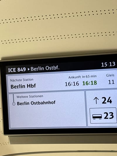 The image shows a digital display inside a train, likely a Deutsche Bahn ICE. The display shows train information, including the train number "ICE 849" heading towards "Berlin Hbf" and "Berlin Ostbahnhof." The current time is 15:13. The next station is "Berlin Hbf," with an arrival time of 16:16, and the platform is "11." "Berlin Ostbahnhof" is listed as a further station. To the right, there's information about track numbers, "24" and "23" with a train icon. The display is mounted on a light-colored interior wall of the train, with a pattern of small dots visible above the screen. The scene is indoors, in what appears to be the passenger area of a modern train. The image does not provide any specific information about the city of Wolfsburg, Germany.