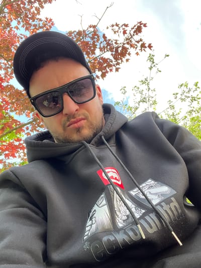 A low-angle shot captures a person wearing a black hoodie with white and gray graphics and a red emblem. The person also wears a black baseball cap and black-framed sunglasses. The face is visible with a short beard and mustache. Behind the person, there are tree branches with green and reddish-brown leaves and patches of blue sky and white clouds. There is no indication of this image being taken in Westerkappeln, Germany.