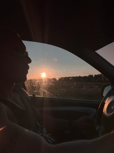 The image shows a person driving a car during sunset, with the sun casting a warm glow. The person is wearing glasses and a tank top, and their arm is resting on the steering wheel. The car appears to be on a road, with trees visible in the background. The overall impression is one of a peaceful and reflective moment, possibly during a road trip. The image is taken from inside the car, looking out the side window. The sky is a gradient of orange and blue, with the sun setting in the distance. The car's interior is dimly lit, with the sun's rays creating a strong silhouette effect. The scene suggests an outdoor setting, possibly a rural or suburban area, given the presence of trees and open space.