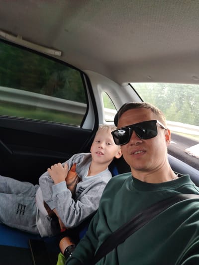 The image shows a selfie taken from inside a car. A man wearing dark sunglasses and a green sweater with a black seatbelt across his chest is in the foreground. Behind him, a boy with blond hair is sitting in the backseat, wearing a gray hoodie and pants. The boy is holding a stuffed animal. The car is moving, as suggested by the blurred scenery outside the window. Based on the geolocation Неволино, Russia, the photo was likely taken within the vicinity of that location.