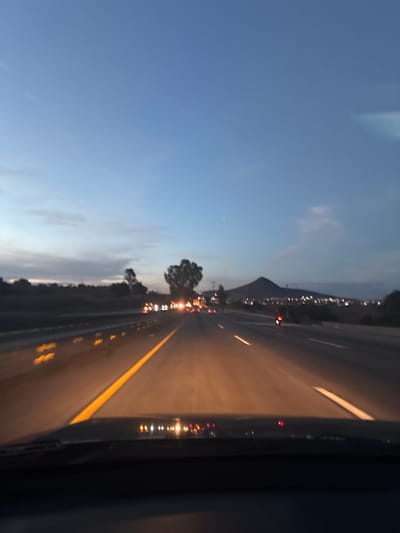 A low-angle view from inside a vehicle moving on a highway at dusk. A yellow line divides the lanes of the highway. Reflections from the vehicle's interior are visible. In the distance, there's a mountain silhouette against a darkening sky. City lights are scattered along the horizon. Approaching traffic creates blurred light trails. Trees are visible along the highway's edge under a blue sky.