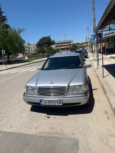 A silver Mercedes-Benz C-Class sedan is parked on the right side of a paved road in Dragoevo, Bulgaria. The car is facing forward, with its license plate reading "CB 8992 KM" visible. The vehicle is positioned next to a sidewalk with a few people visible in the background, along with buildings and trees. The sky is clear and blue, indicating a sunny day. The scene appears to be a street in a town or city, possibly a residential or commercial area. There is a pedestrian crossing visible on the left side of the road.