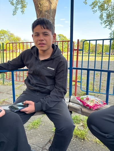 The image depicts an outdoor scene in Glava, Bulgaria, likely a park or playground, on a bright, sunny day with a clear blue sky. The primary subject is a young person, possibly male, with short dark hair, who is sitting on a wooden bench and looking directly at the camera with a slight smile, conveying a relaxed demeanor. They are dressed in a black zipped hoodie with the word "STARS" printed in white on the left chest, and matching black sweatpants.
Two other individuals are partially visible. To the right of the main subject, another person is seated on the same bench, with only their black-clad leg and part of their torso visible. Next to them, resting on the bench, is a colorful snack bag, possibly containing chips or nuts, with Cyrillic text partially readable as "БЯЛА," "САЛА," and "ЦАРСКА," which likely refers to a snack brand such as "Белла Царска салата." In the lower right foreground, the legs of a third person are visible, also wearing black pants, holding a smartphone.
The background features prominent blue and red metal railings, suggesting playground equipment or a fence. A large tree trunk is visible on the far left. The ground is a mixture of green grass, scattered vegetation, and a light-colored paved surface. The time of day appears to be midday or afternoon due to the strong sunlight and clear sky, suggesting pleasant weather for outdoor relaxation.