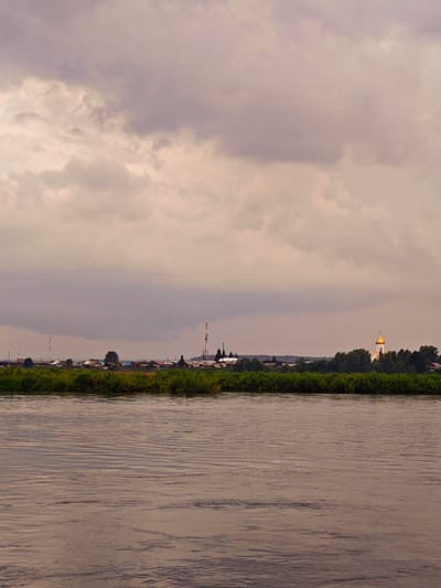 A wide, calm river fills the foreground of the image, its surface rippled by a gentle breeze. The water has a muted, brownish hue, reflecting the overcast sky. Beyond the river, a lush green embankment is covered in dense foliage. Nestled behind the vegetation is a small village. The buildings are a mix of houses with colorful roofs and some industrial structures, including two tall communication towers. To the right of the village, a golden-domed church stands out against the greenery, its spire catching the light. The sky above is filled with heavy, dark clouds, creating a somber, atmospheric mood. The overall scene suggests a quiet, rural setting, likely in the town of Kazachinskoe, Russia, given the architectural style of the church and the general landscape. No people are visible in the image.